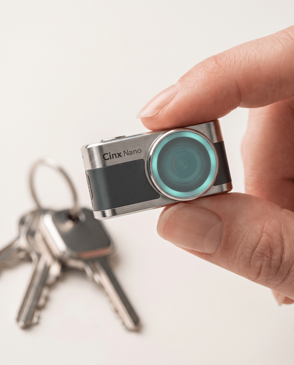 The Cinx Nano camera held between fingertips beside keys, showing its tiny actual size